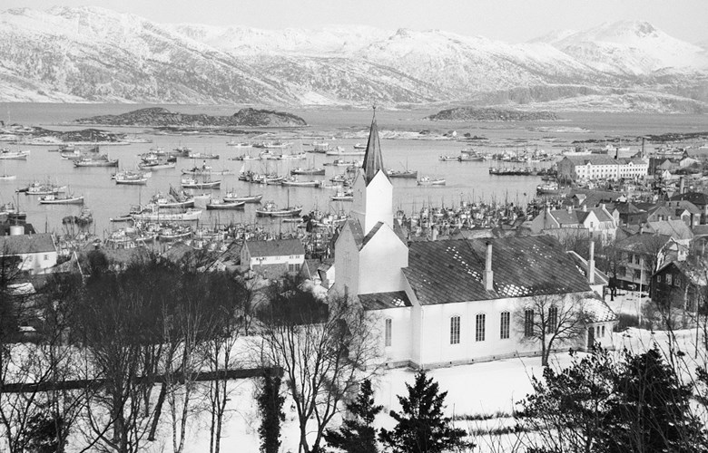 Utsyn frå Likkjeåsen og nord-austover, med kyrkja i framgrunnen, byen og fjella i nordaust. Fiskarflåten ligg i hamn, februar 1953.
