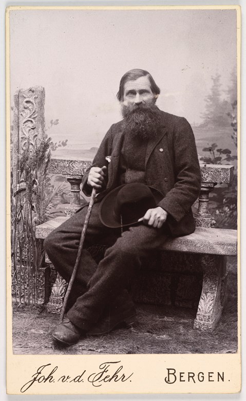 Bilete av ein mann som sit på ein benk, kledd i svart, med skjegg, og med ein vandrestav i ei hand og ein hatt i den andre.