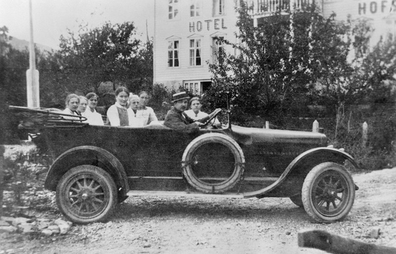 Dette er den første turbilen i Indre Sogn Automobilselskap A/S,  ein Chandler med sju seter, kjøpt i Bergen i juni 1916. Sjåføren er Anton Andersen som og var bilmekanikar. Han var såkalla garantisjåfør den første tida. Ingen andre kunne køyra bil i Sogndal. Passasjerane er truleg jenter frå Sogndal. Fartsgrensa var 30 km/t, og selskapet måtte stilla garanti for mogeleg skade før køyringa tok til, sjølv om det ikkje var sjåføren si skuld.

 