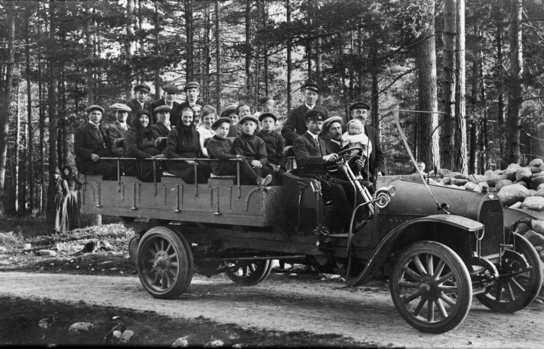 Dette var den første bilen A/S Fjordenes Automobilselskap kjøpte i 1913. Passasjerane er på veg til Breim, der dei skulle ta motorbåten inn til Førde. 21 personar sit på lasteplanet. Dei første bilane var små lastebilar. Når dei vart nytta til persontransport, vart det sett benkar eller stolar på lasteplanet. Olai Farsund, frå Førde, var sjåfør. Han var ein av dei dei første sjåførane i selskapet. 

 