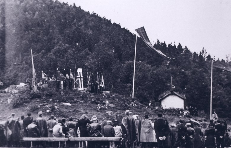 Frå avdukingshøgtida ved ferjestaden Våge søndag, 15. august 1943. Bautasteinen er sveipt i det norske flagget og omkransa av flagg og banner. Heimestyrkar øydela minnesmerket i juni 1945.
