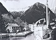 Slik har mange reisande møtt Balestrand når dei har kome med båt inn fjorden. Her kjem D/S <i>Kommandøren</i> til kai i Balestrand, truleg på utgåande rute kl. 12 på dagen, ein sommardag i 1936. <i>Kommandøren</i> gjekk i rute mellom Bergen og Indre Sogn. Her ser vi bryggja som vart  bygt i 1911 og Kaihuset. Ved sida av kan ein så vidt skimte bygningen Bom Bom før Cooperhuset.