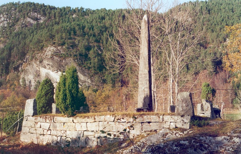 Minnebautaen over Eidsvoll-mannen Ole Elias Holck er reist på ei høgd like ved gardstunet i Alværa. Han er fundamentert i berget og ruvar kring 5 meter i været. På den oppmura foten rundt, er sett steinar i kvart hjørne. Hovudinnskrifta er på på den sørvende sida mot fjorden.